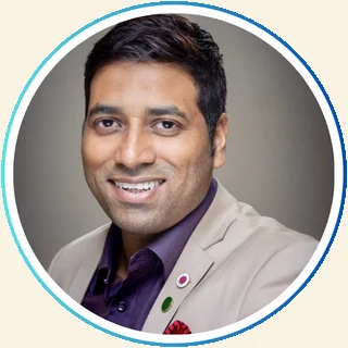 Asslam's LinkedIn profile photo: a studio headshot on a plain grey background, face filling the circular crop, open smile and direct eye contact, cream blazer and lapel pins